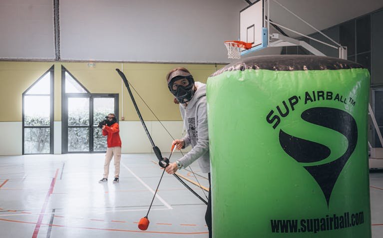 Exciting indoor archery tag game with players in protective gear, aiming and hiding behind barriers.